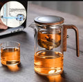 Glass teapot with wooden handle and a small glass cup on a wooden surface.