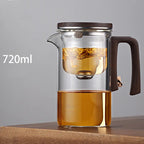 Clear glass teapot with wooden handle and lid, filled with tea, on a wooden surface.