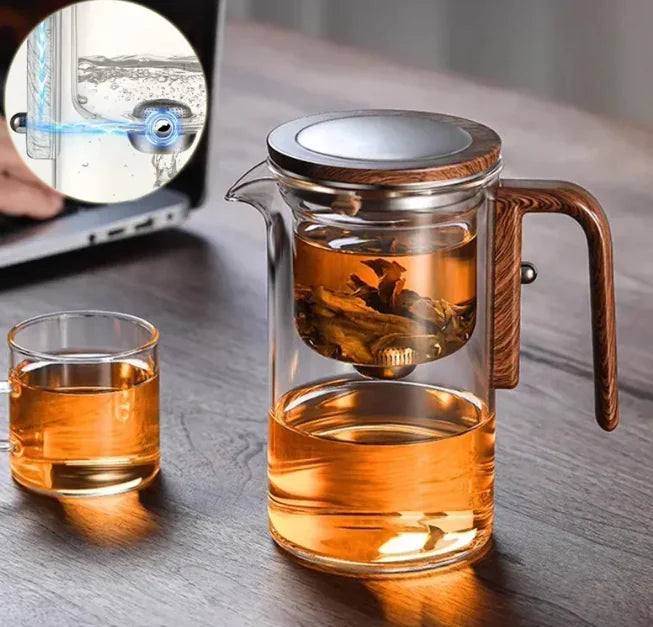 Glass teapot with wooden handle and a smaller glass cup on a wooden surface, with a laptop in the background.
